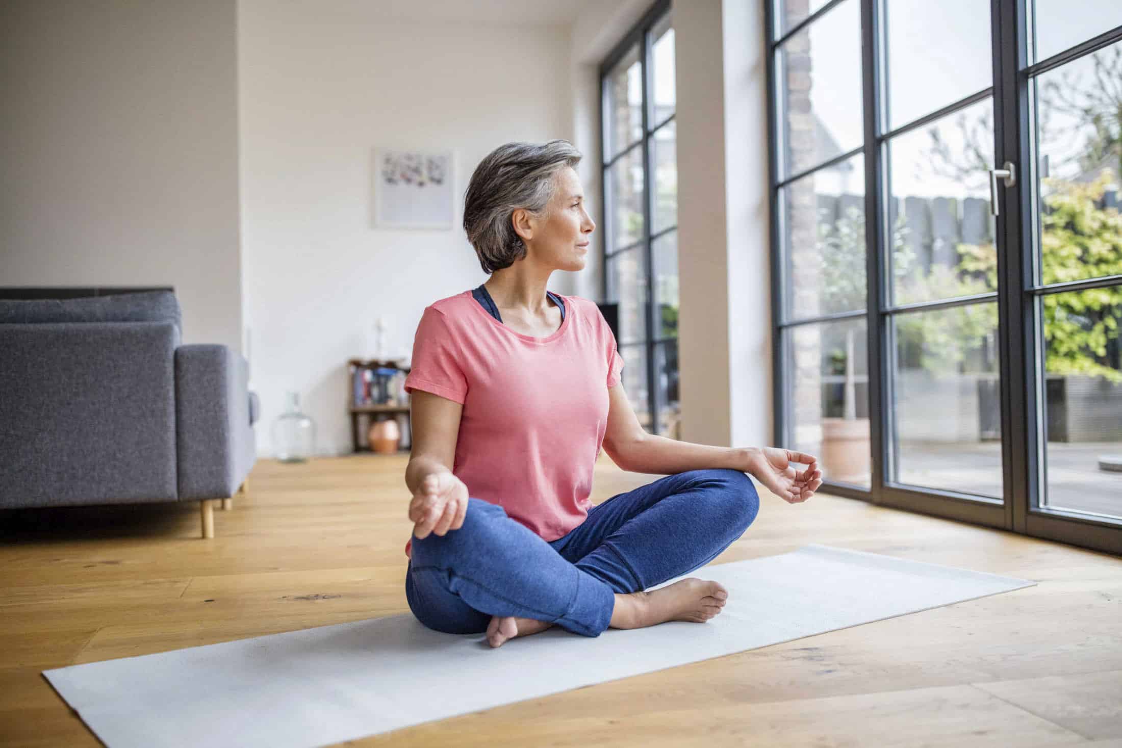 Eine Frau mit kurzen grauen Haaren sitzt im Schneidersitz auf einer Yogamatte in einem hellen Wohnzimmer und meditiert friedlich, während sie aus großen Fenstern schaut. Sie trägt ein rosa Oberteil und eine blaue Hose. © Fotografie Tomas Rodriguez