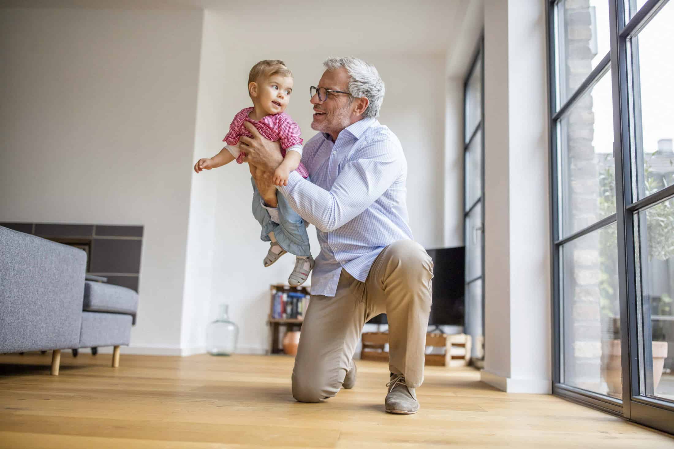 In einem hellen, modernen Wohnzimmer mit großen Fenstern kniet ein älterer Mann auf dem Holzboden und hebt spielerisch ein lächelndes kleines Kind hoch. © Fotografie Tomas Rodriguez