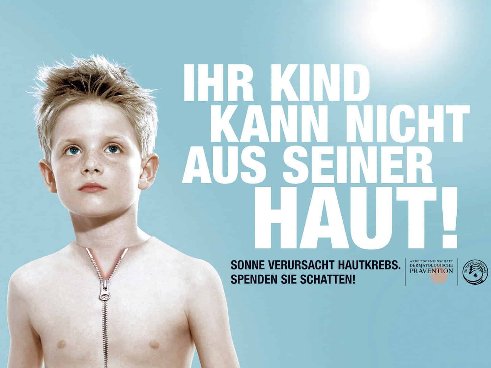 Ein kleiner Junge mit heller Haut und blondem Haar steht vor einem blauen Hintergrund. Auf seiner Brust befindet sich ein großer, teilweise geöffneter Reißverschluss, unter dem blasse Haut zum Vorschein kommt. Ein großer deutscher Text warnt vor Hautkrebs und Sonnenschutz für Kinder. © Fotografie Tomas Rodriguez