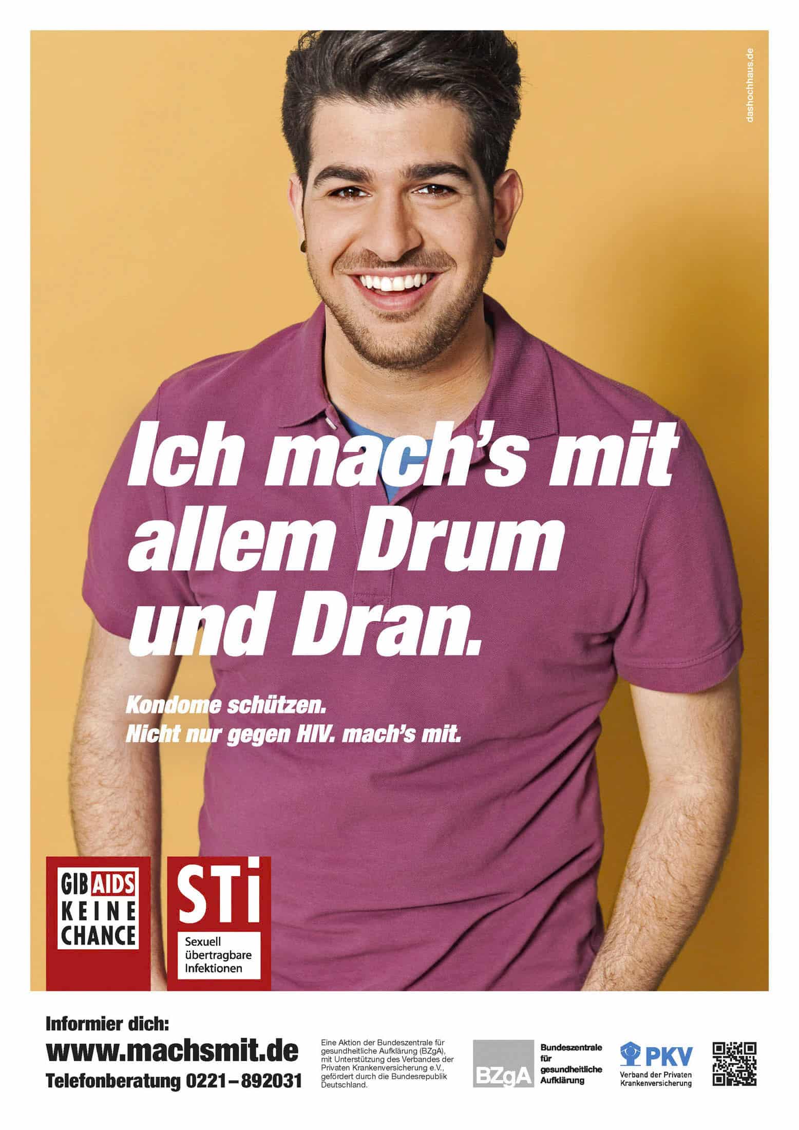 Ein lächelnder junger Mann in einem rosa Poloshirt steht vor einem gelben Hintergrund. In großer weißer Schrift steht: "Ich mach's mit allem Drum und Dran." Das Plakat wirbt für die HIV- und STI-Prävention, die Verwendung von Kondomen und das Bewusstsein für sexuelle Gesundheit. © Fotografie Tomas Rodriguez