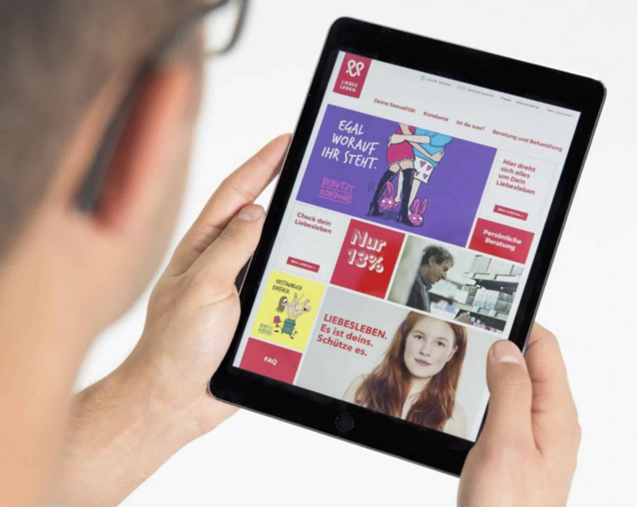 Eine Person hält ein Tablet in der Hand, auf dem eine deutschsprachige Website mit bunten Bannern, Fotos von Menschen und gesundheitsbezogenen Botschaften zu den Themen Liebe, Beziehungen und sichere Praktiken angezeigt wird. © Fotografie Tomas Rodriguez