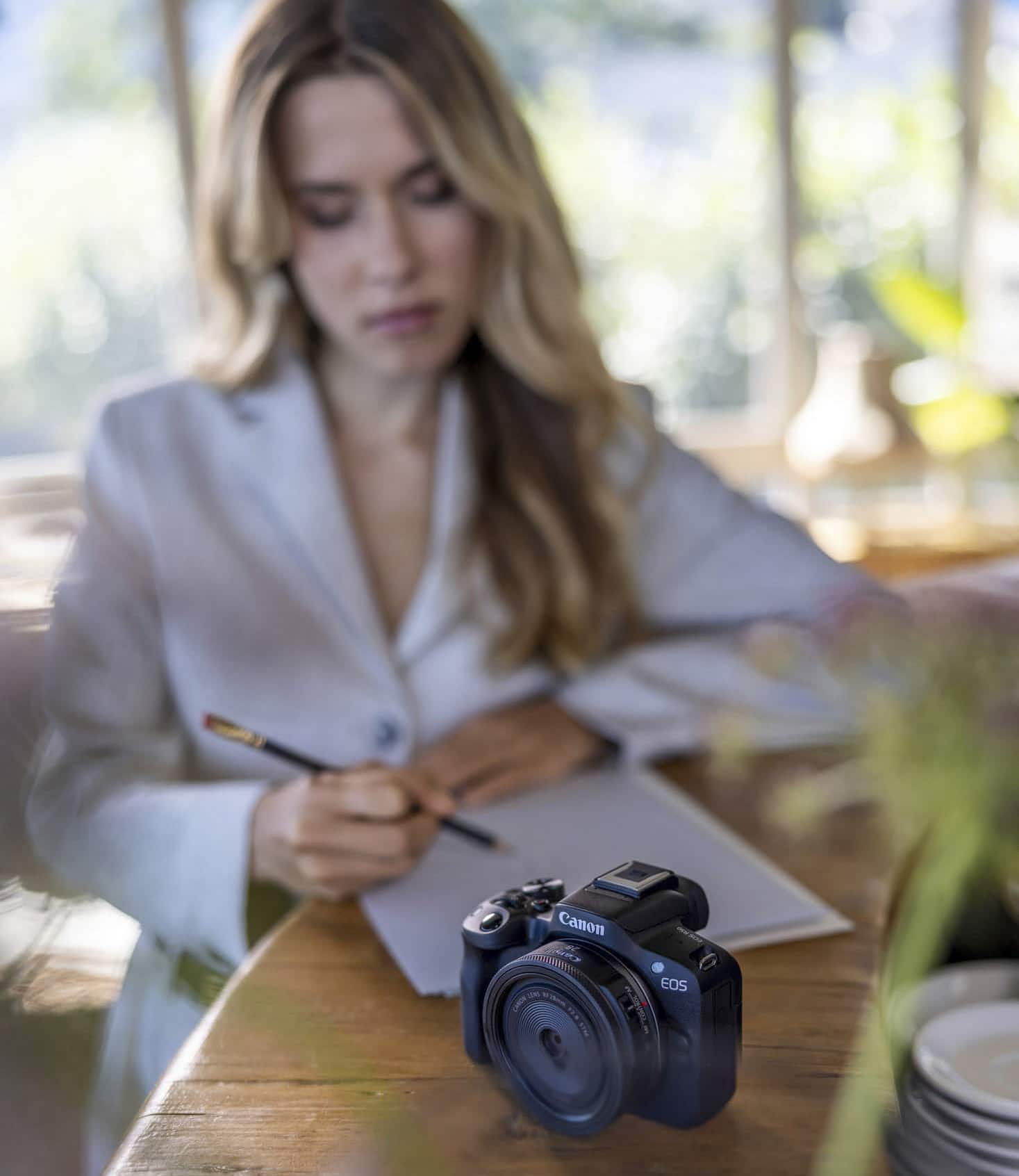Eine Frau mit langen Haaren sitzt an einem Holztisch und schreibt mit einem Bleistift in ein Notizbuch. Eine Canon EOS-Kamera ist auf den Tisch im Vordergrund fokussiert. Die Szene ist sanft mit natürlichem Licht beleuchtet. © Fotografie Tomas Rodriguez