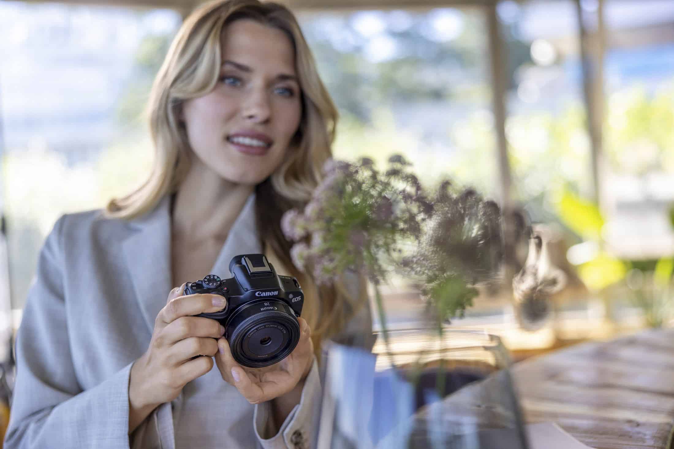 Eine Frau, die eine Canon-Kamera in der Hand hält, lächelt sanft in einem Innenraum mit verschwommenen lila Blumen im Vordergrund und natürlichem Licht, das durch große Fenster strömt. © Fotografie Tomas Rodriguez