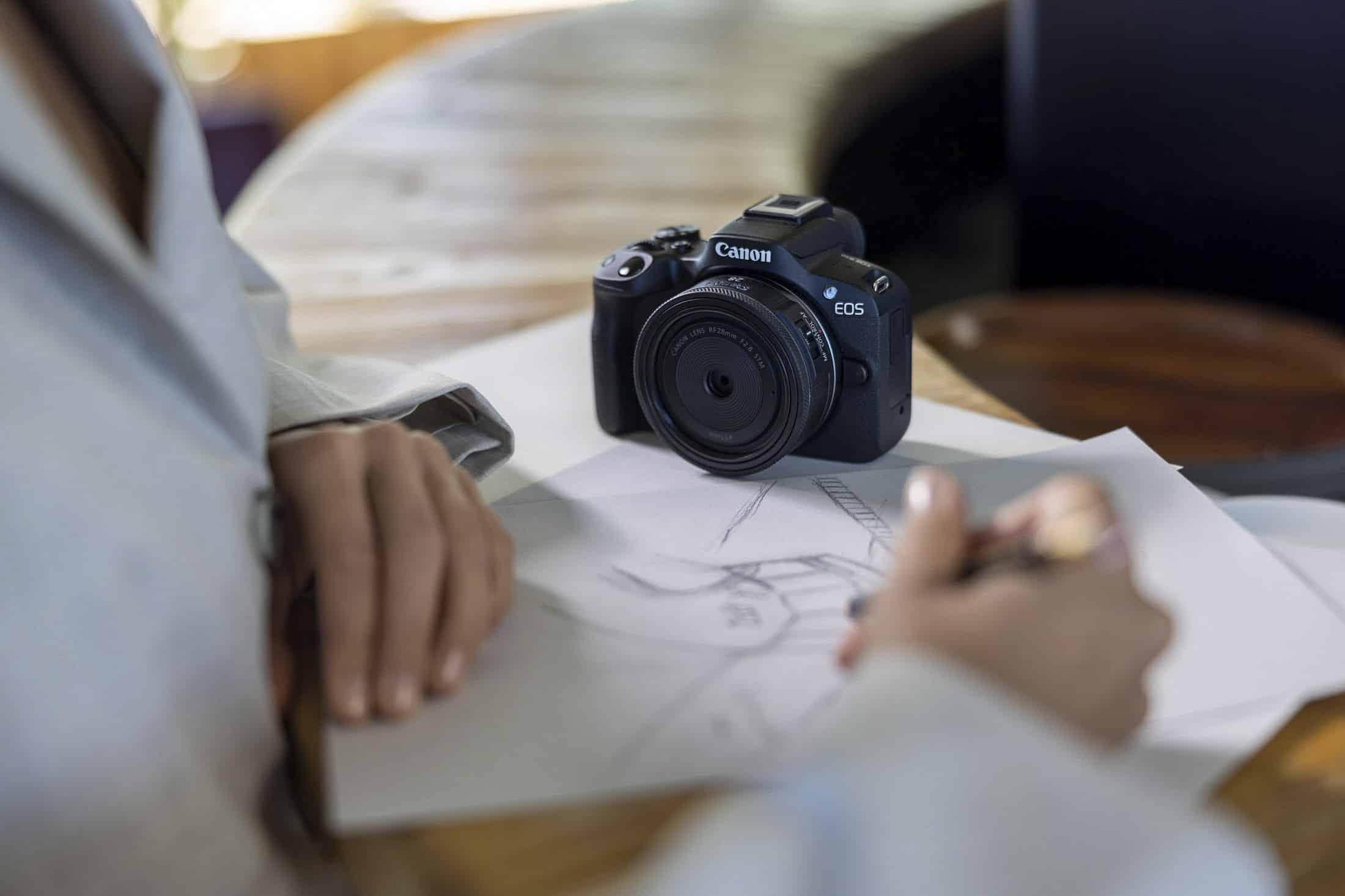 Eine Person, die an einem Holztisch auf weißem Papier skizziert, während in der Nähe eine Canon EOS-Kamera ruht. Der Fokus liegt auf der Kamera und den Händen der Person. © Fotografie Tomas Rodriguez