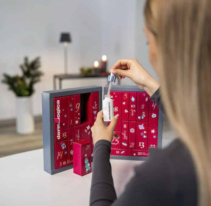 Eine Person öffnet einen roten und grauen Adventskalender auf einem Tisch und nimmt ein kleines Fläschchen mit einer Pipette mit der Aufschrift "Dermalogica" heraus. Im Hintergrund sind unscharf Kerzen und eine Pflanze zu sehen. © Fotografie Tomas Rodriguez