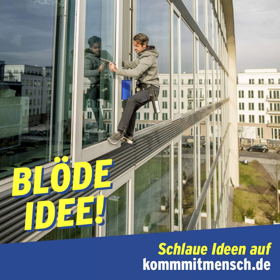 Ein Fensterputzer sitzt gefährlich auf einem schmalen Sims vor einem hohen Glasgebäude. Der Text in Deutsch lautet: "BLÖDE IDEE!" (Dumme Idee!) und "Schlaue Ideen auf kommmitmensch.de" (Schlaue Ideen auf kommmitmensch.de). © Fotografie Tomas Rodriguez