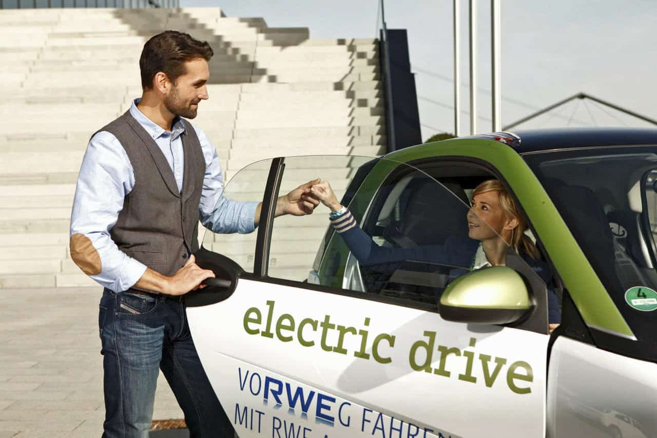 Ein Mann steht vor einem Elektroauto mit der Aufschrift "electric drive" und hält die Hand einer lächelnden Frau, die darin sitzt. Im Hintergrund sind moderne Treppen und ein Gebäude zu sehen. © Fotografie Tomas Rodriguez