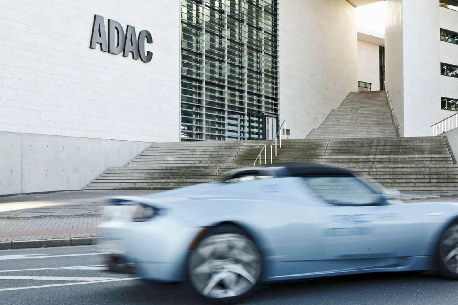 Ein hellblauer Sportwagen fährt an einem modernen Gebäude mit großen Glasfenstern und dem Schriftzug "ADAC" an der Außenwand neben einer breiten Treppe vorbei. © Fotografie Tomas Rodriguez