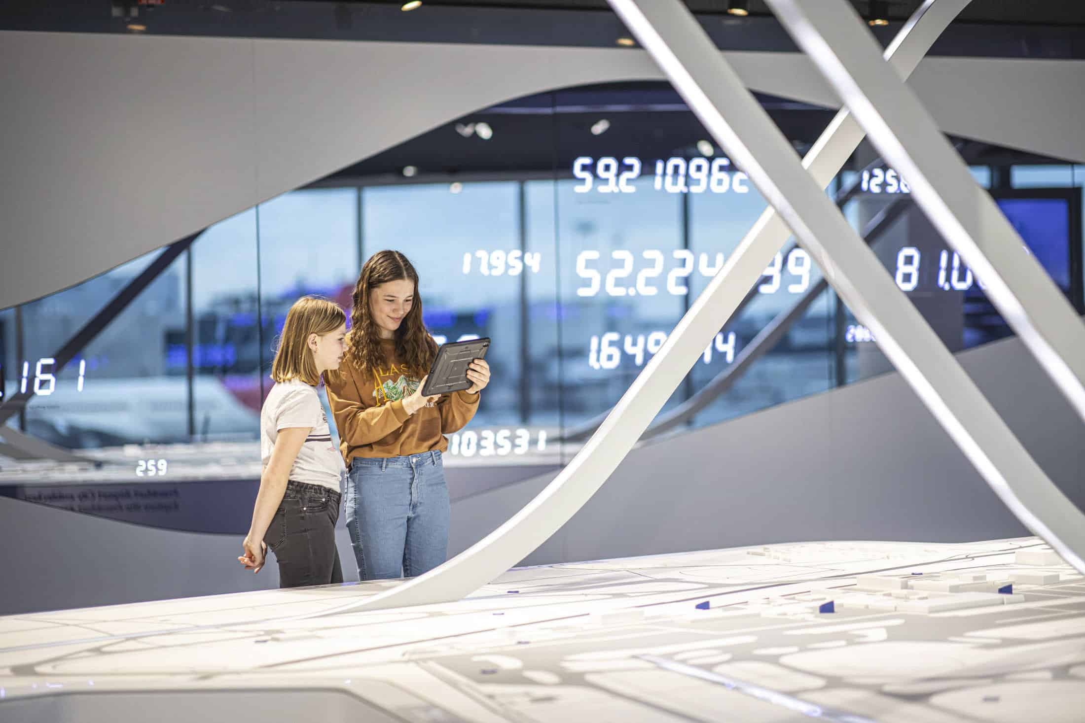 Zwei Mädchen stehen zusammen in einer modernen High-Tech-Ausstellung und schauen auf ein Tablet. An einer Wand hinter ihnen sind große digitale Zahlen zu sehen, und abstrakte weiße Strukturen sind Teil der Installation. © Fotografie Tomas Rodriguez