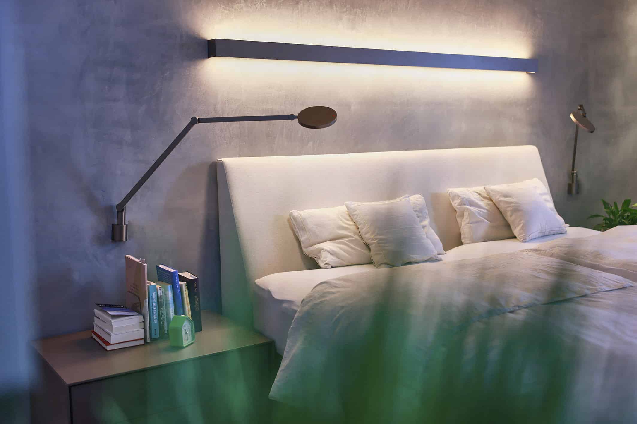 Modernes Schlafzimmer mit einer stilvollen Leuchte über einem weißen Polsterbett, drei Kissen, einem Nachttisch mit gestapelten Büchern und einer an der Wand befestigten Leselampe; weiches, grün getöntes Licht im Vordergrund. © Fotografie Tomas Rodriguez