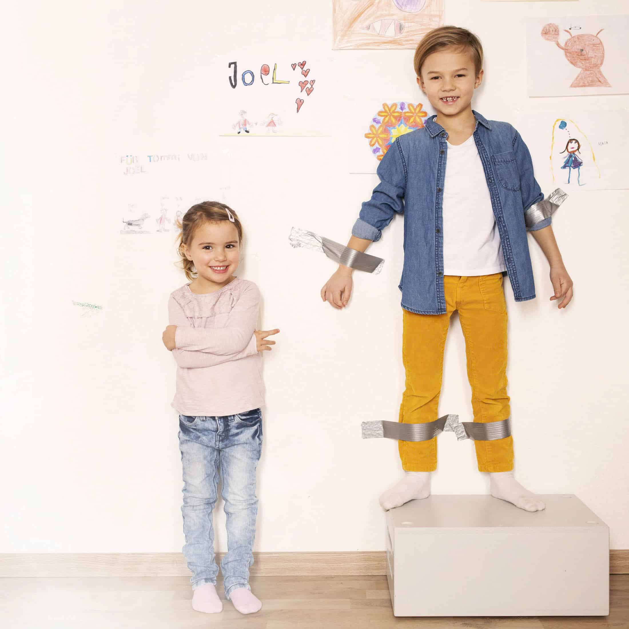 Ein Junge ist mit Klebeband an eine Wand geklebt, steht auf einer Kiste und lächelt. Ein junges Mädchen steht mit verschränkten Armen neben ihm und lächelt. An der Wand hinter ihnen sind Kinderzeichnungen zu sehen. © Fotografie Tomas Rodriguez
