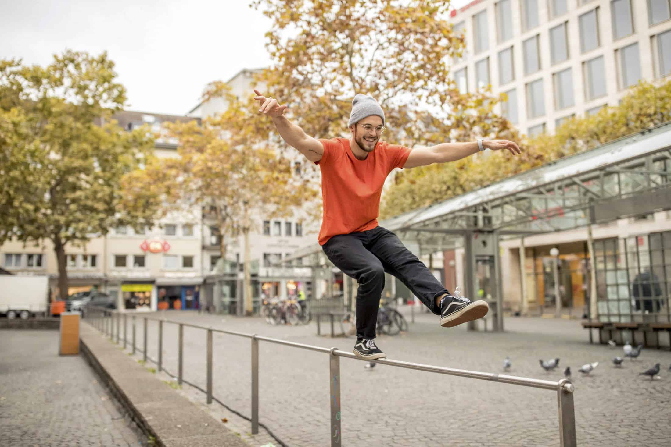 Eine lächelnde Person in einem roten Hemd und einer grauen Mütze balanciert spielerisch auf einem Metallgeländer auf einem städtischen Platz, mit Herbstbäumen, Gebäuden und Tauben im Hintergrund. © Fotografie Tomas Rodriguez
