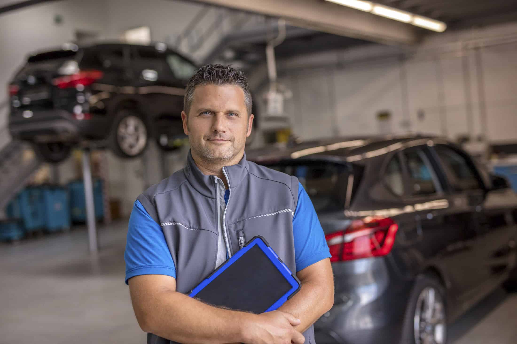 Ein Mann in einem blauen Hemd und einer grauen Weste steht in einer Autowerkstatt und hält ein Tablet in der Hand. Hinter ihm stehen zwei Autos, eines auf einer Hebebühne, das andere auf dem Boden der Werkstatt geparkt. Die Umgebung wirkt hell und aufgeräumt. © Fotografie Tomas Rodriguez