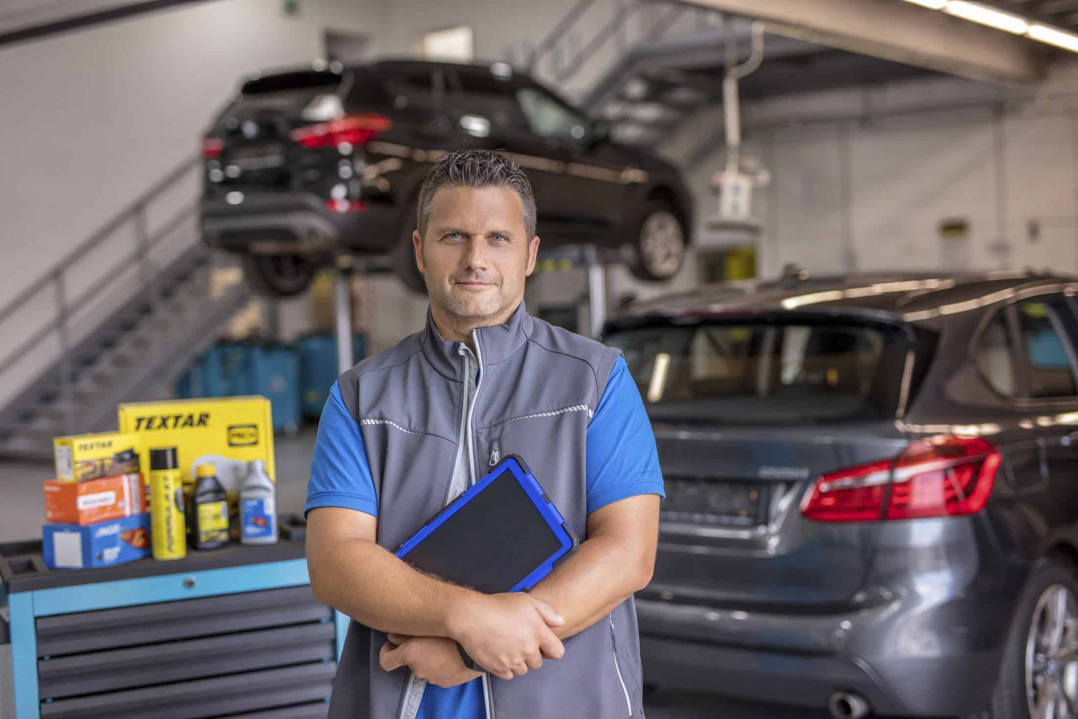 Ein Mechaniker mit einem Klemmbrett steht in einer Autowerkstatt. Hinter ihm stehen zwei dunkle Autos - eines auf einer Hebebühne und eines auf dem Boden. Auf einem Wagen in der Nähe sind Kfz-Werkzeuge und Zubehör zu sehen. © Fotografie Tomas Rodriguez