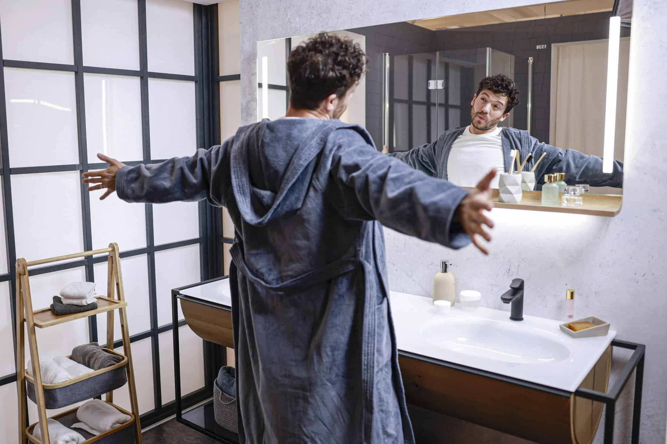 Ein Mann im Bademantel steht mit ausgestreckten Armen in einem modernen Badezimmer und betrachtet sich im Spiegel über dem Waschbecken. In dem Raum sind Toilettenartikel und Handtücher auf einem Holzregal ordentlich angeordnet. © Fotografie Tomas Rodriguez