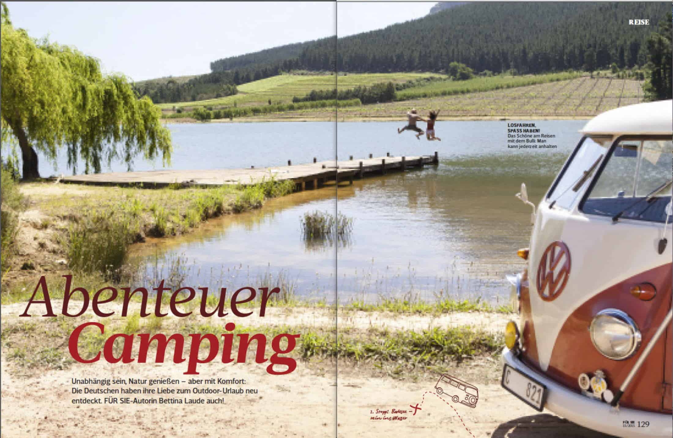 Ein alter VW-Wohnwagen steht an einem See mit bewaldeten Hügeln im Hintergrund. Zwei Personen springen von einem Holzsteg ins Wasser und genießen einen sonnigen Tag in der Natur. Der Text lautet "Abenteuer Camping. © Fotografie Tomas Rodriguez
