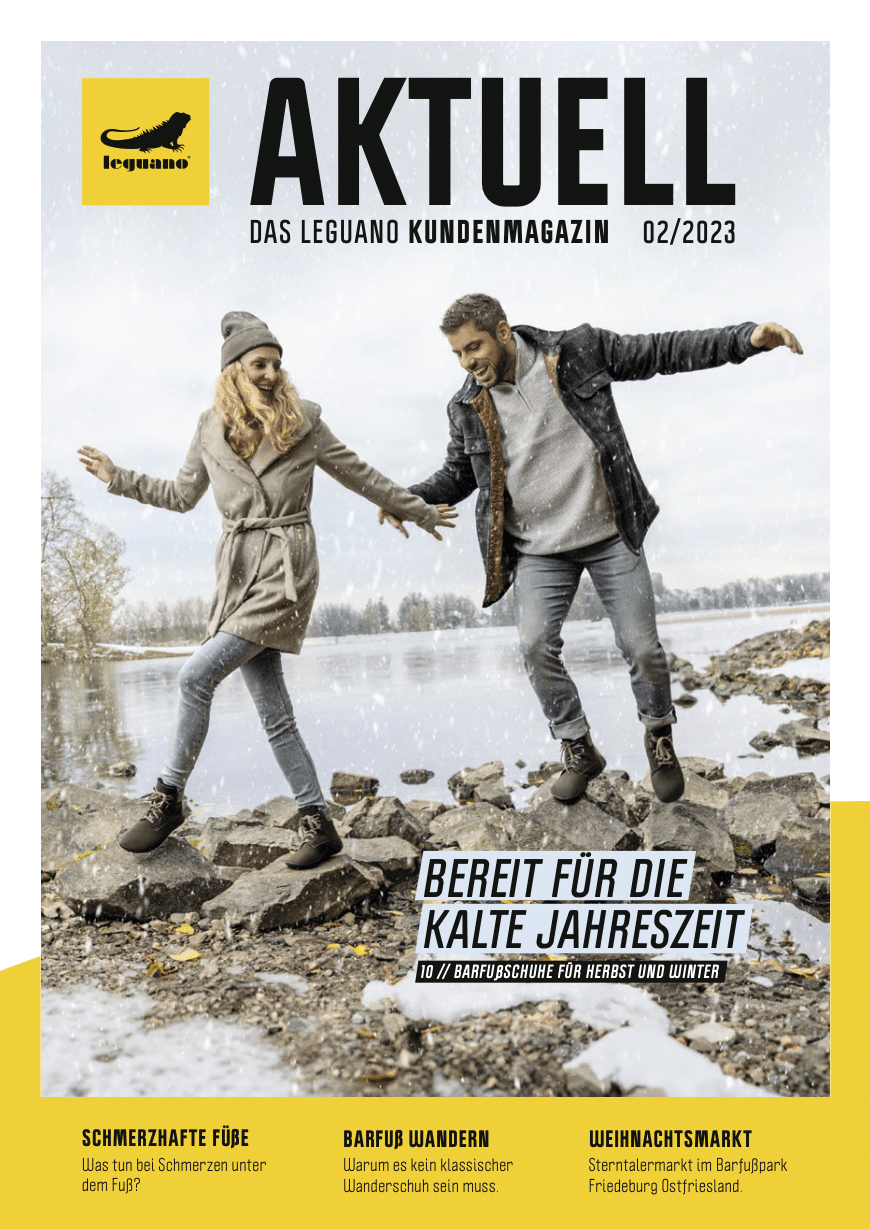 Eine lächelnde Frau und ein lächelnder Mann in herbstlicher Kleidung balancieren fröhlich auf einem verschneiten Baumstamm im Freien. Sie sind auf der Titelseite des "Leguano Kundenmagazins" mit Artikeln über Barfußschuhe für die kälteren Jahreszeiten abgebildet. © Fotografie Tomas Rodriguez