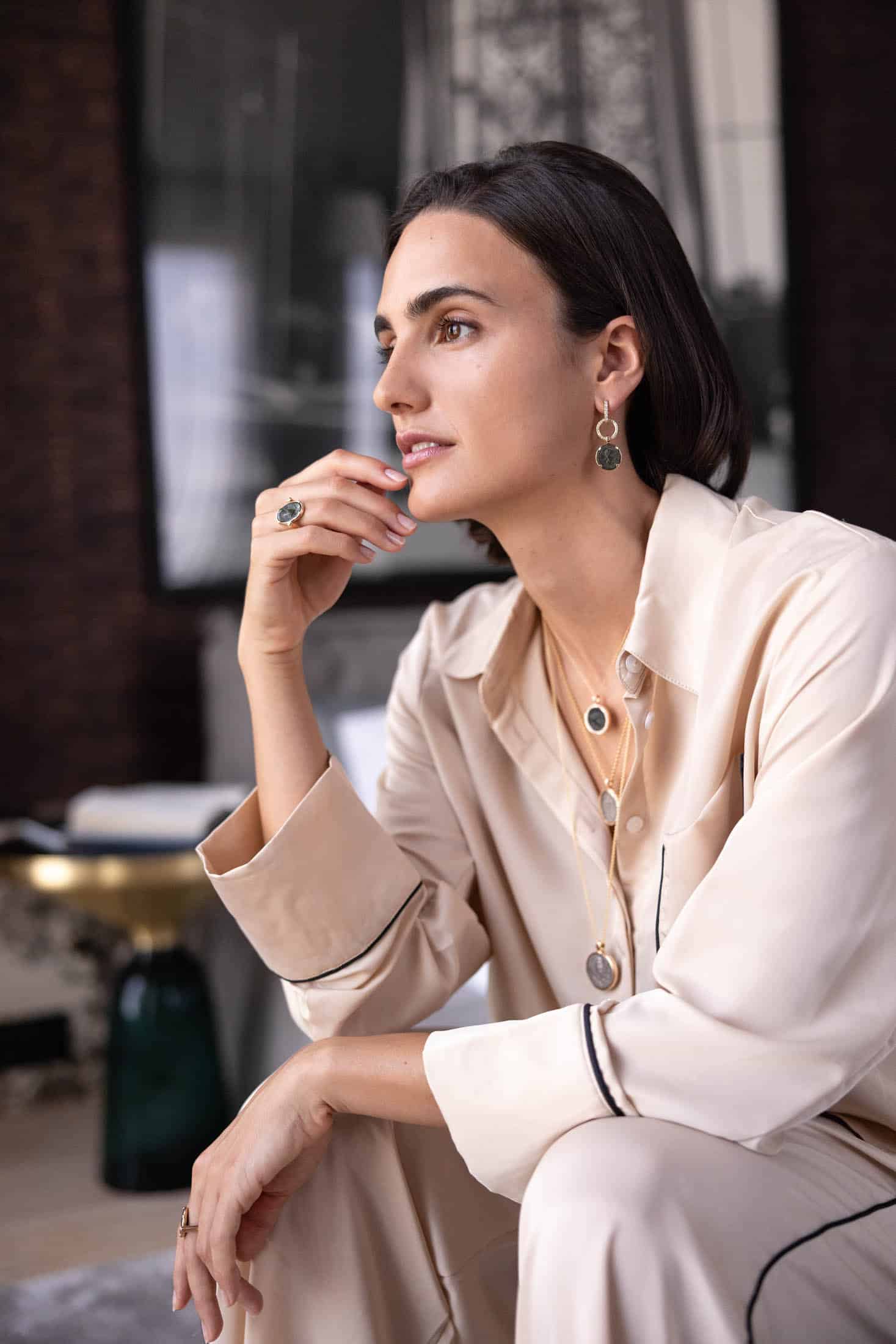 Eine Frau mit kurzen, dunklen Haaren sitzt nachdenklich da. Sie trägt eine cremefarbene Satinbluse und mehrreihigen Schmuck, darunter Ringe, Ohrringe und Halsketten mit Steinen. In einem sanft beleuchteten, eleganten Raum blickt sie zur Seite. © Fotografie Tomas Rodriguez