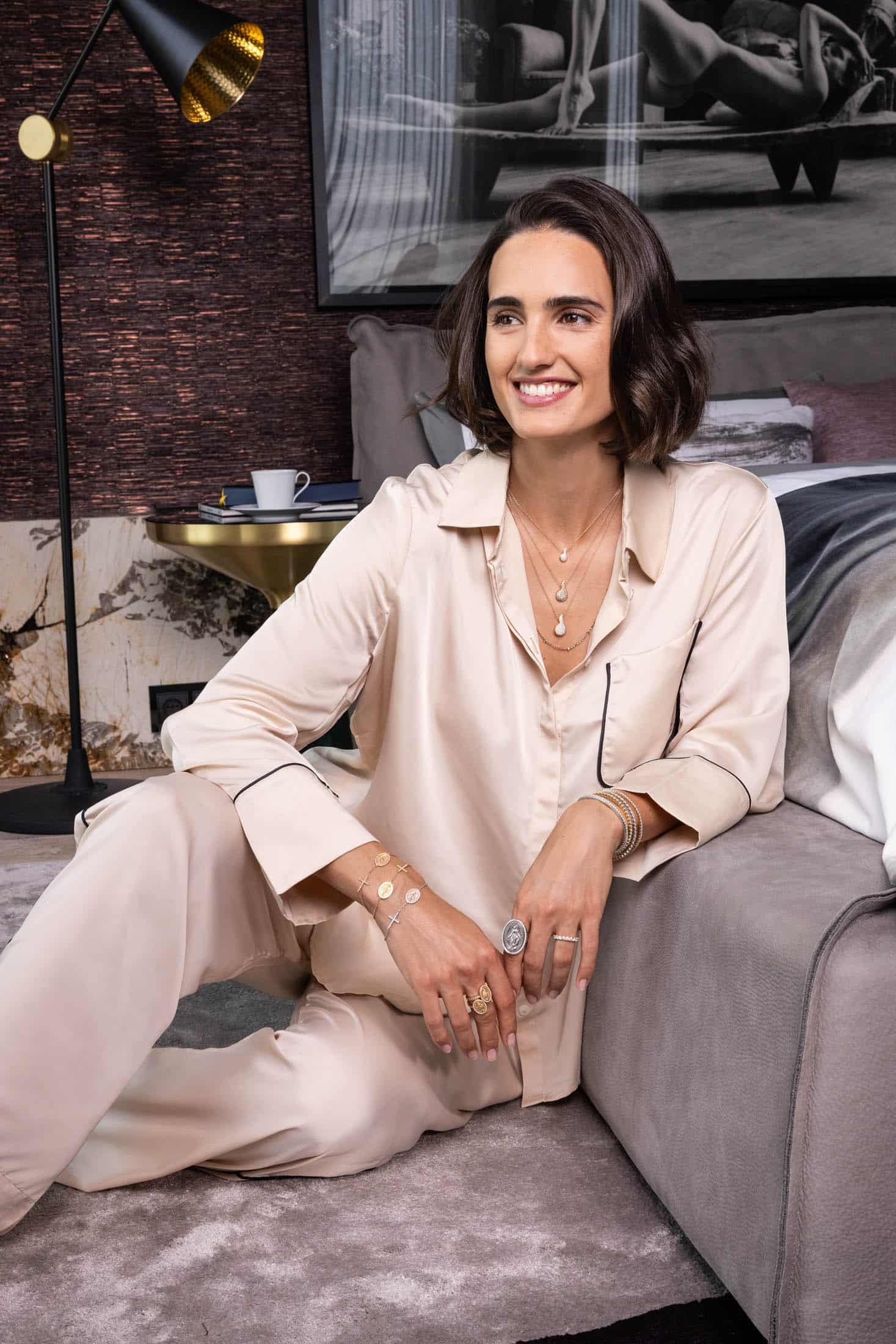 Eine Frau in einem cremefarbenen Seidenpyjama sitzt lächelnd auf dem Boden eines Schlafzimmers. Sie trägt mehrreihigen Goldschmuck. Hinter ihr stehen eine goldene Lampe, ein Beistelltisch mit einer Tasse und ein Bett. An der Wand hängt ein schwarz-weißes Kunstfoto. © Fotografie Tomas Rodriguez