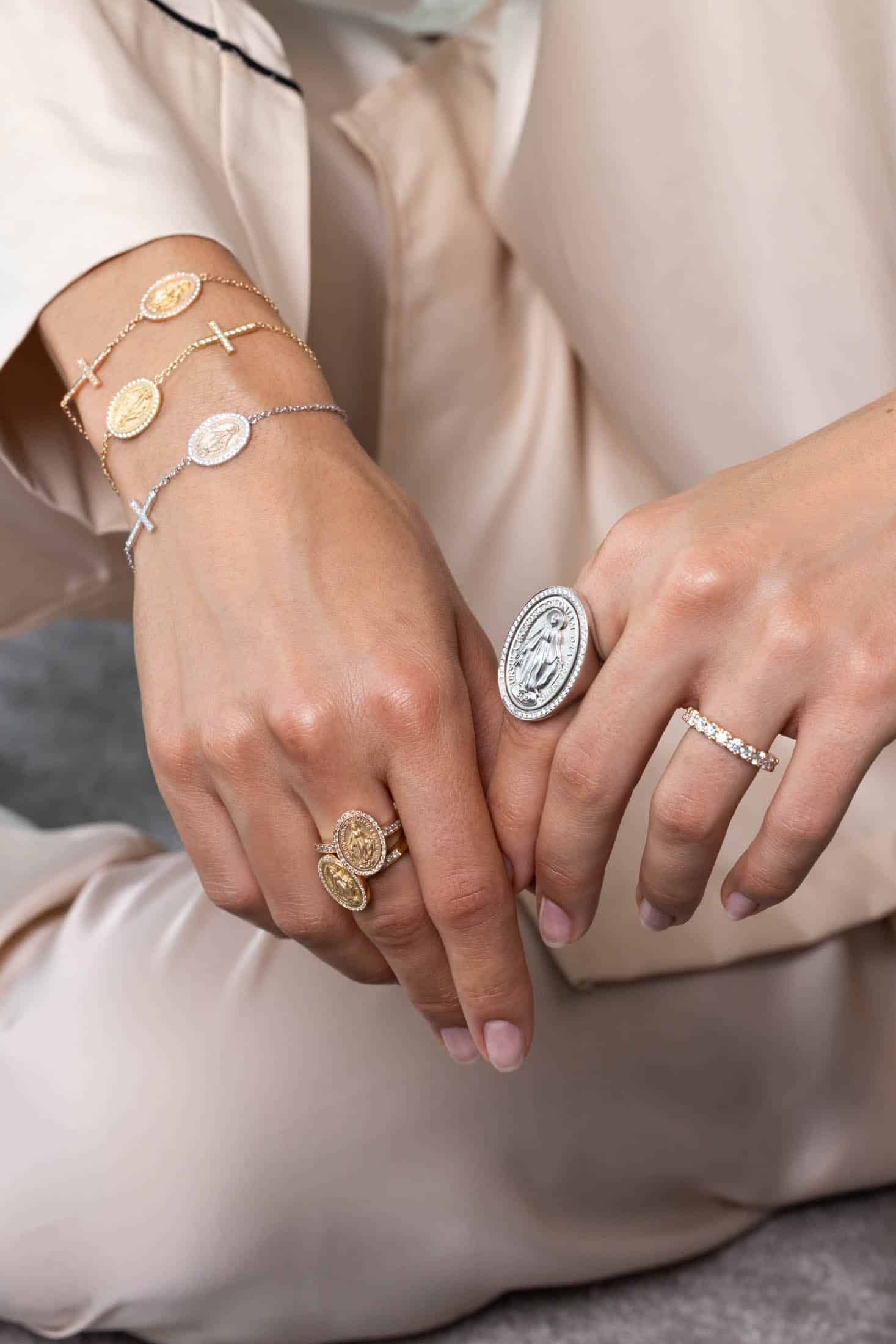 Nahaufnahme von Händen, die Gold- und Silberringe sowie Armbänder mit Münz- und Kreuzmotiven tragen, vor einem neutralen Hintergrund. © Fotografie Tomas Rodriguez