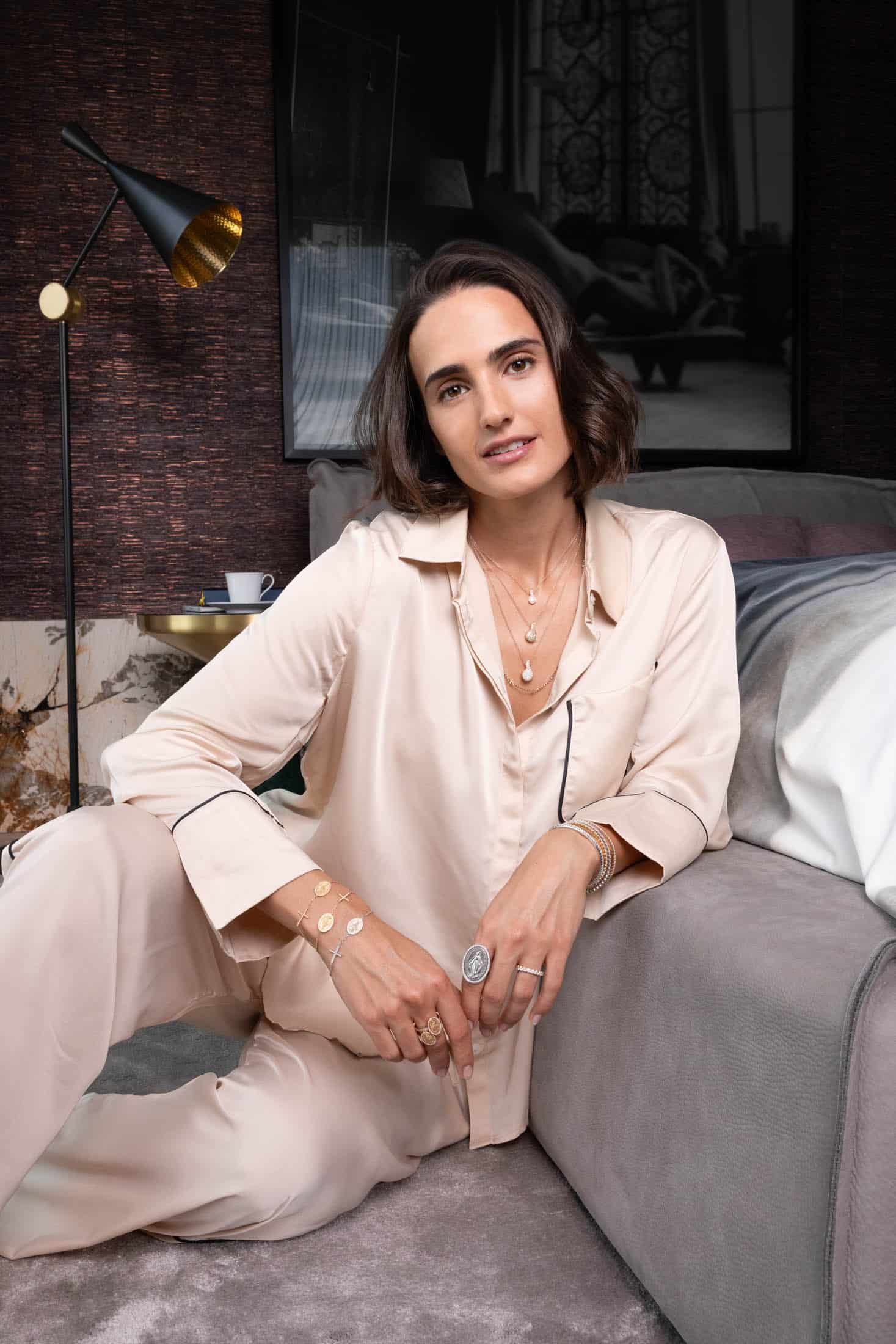 Eine Frau in einem cremefarbenen Satin-Pyjama sitzt auf dem Boden neben einem Sofa und trägt mehrere Halsketten, Ringe und Armbänder übereinander. Sie lächelt leicht, hat kurzes dunkles Haar und befindet sich in einem stilvollen, gemütlichen Interieur mit moderner Einrichtung. © Fotografie Tomas Rodriguez