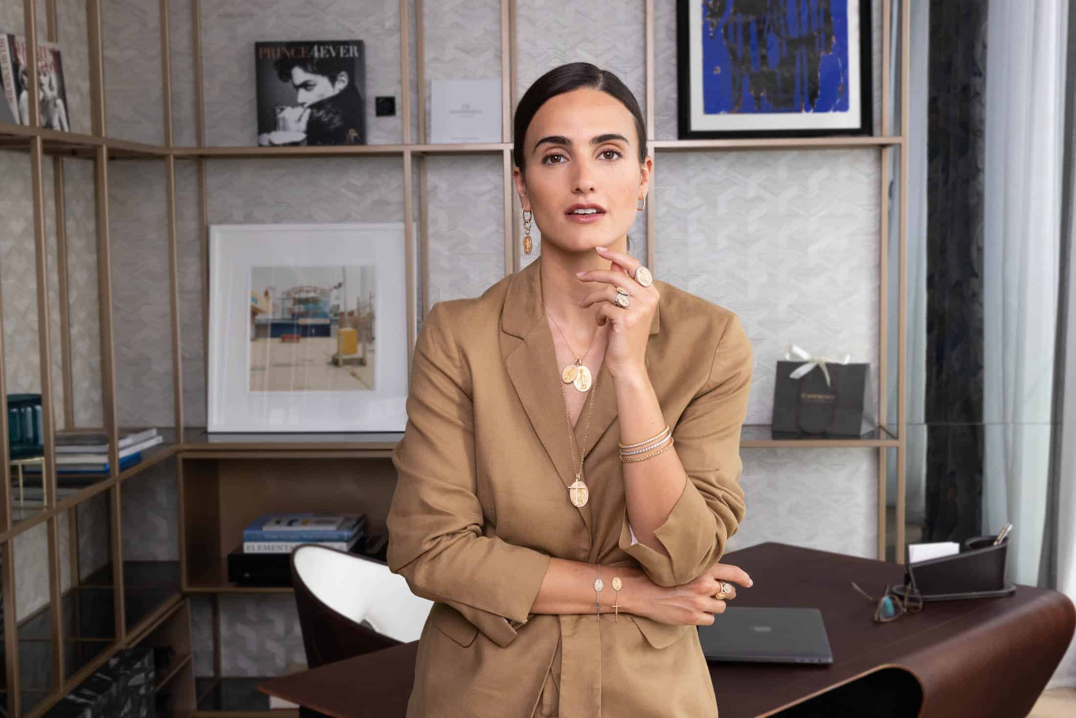 Eine Frau in einem hellbraunen Blazer steht in einem modernen Büro, hinter ihr sind Regale, Bücher und gerahmte Kunstwerke zu sehen. Sie posiert selbstbewusst, trägt Goldschmuck und berührt mit einer Hand ihr Gesicht. © Fotografie Tomas Rodriguez