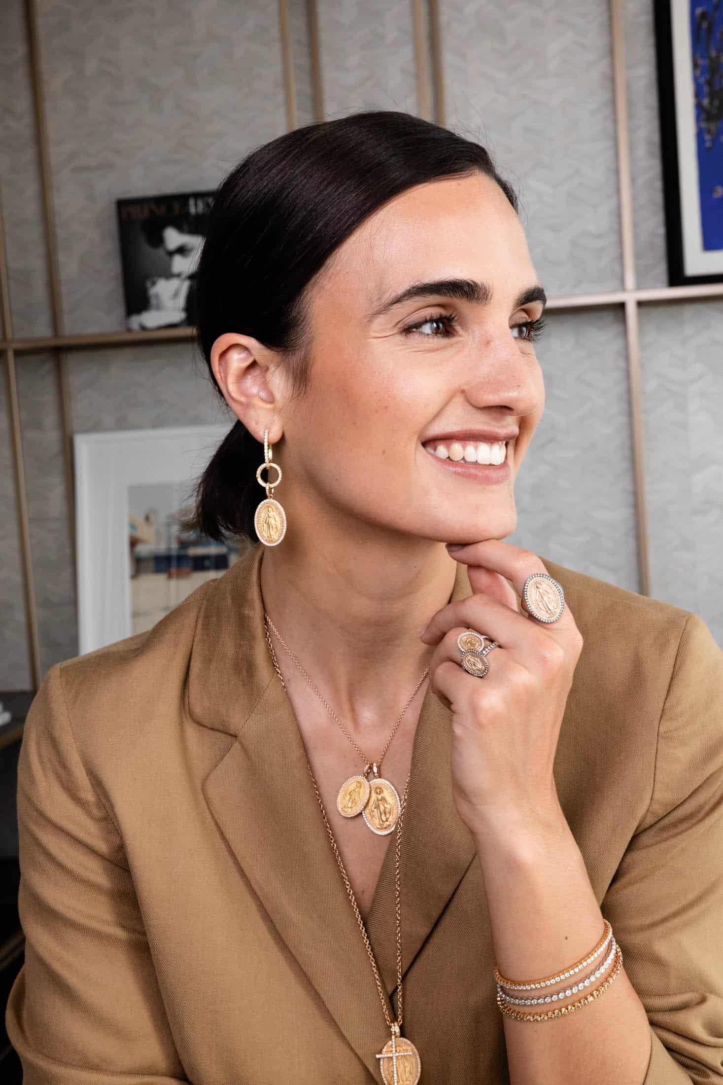 Eine Frau mit dunklem Haar, das zu einem tief sitzenden Dutt zusammengebunden ist, trägt einen hellbraunen Blazer und mehrreihigen Goldschmuck, darunter Ohrringe, Ringe, Armbänder und Halsketten. Sie lächelt und blickt nach rechts. Im Hintergrund sind gerahmte Kunstwerke und eine dekorativ gestaltete Wand zu sehen. © Fotografie Tomas Rodriguez