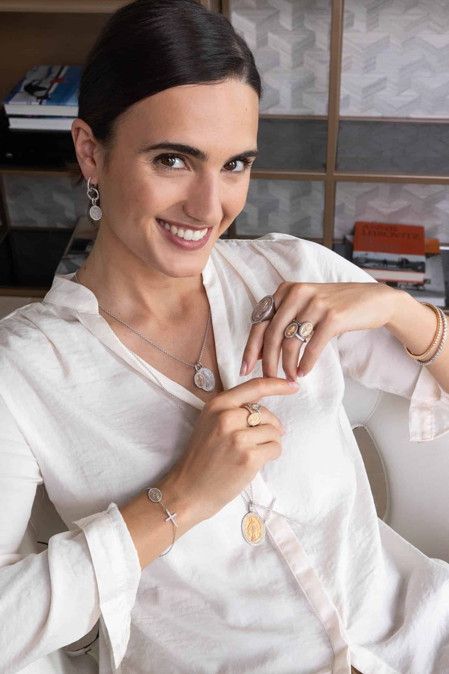 Eine Frau in einer weißen Bluse lächelt, während sie vor einem modernen, mit Bücherregalen gesäumten Hintergrund verschiedene Silber- und Goldschmuckstücke präsentiert, darunter Ringe, Armbänder, Ohrringe und Halsketten. © Fotografie Tomas Rodriguez