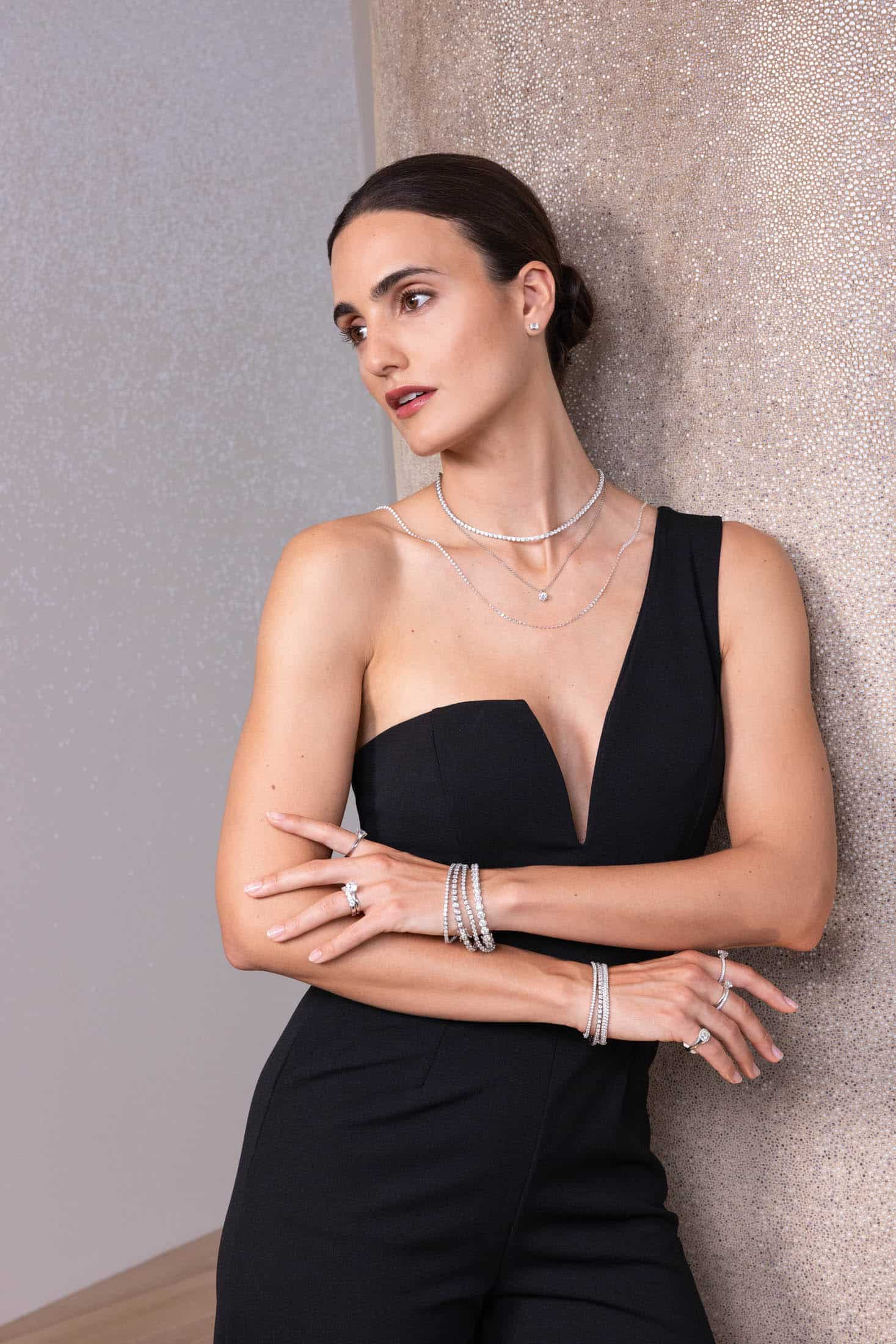 Eine Frau in einem schwarzen, trägerlosen Overall lehnt an einer strukturierten Wand und trägt mehrere Halsketten, Armbänder und Ringe übereinander. Sie blickt mit verschränkten Armen zur Seite und präsentiert dabei ihren eleganten Schmuck. © Fotografie Tomas Rodriguez