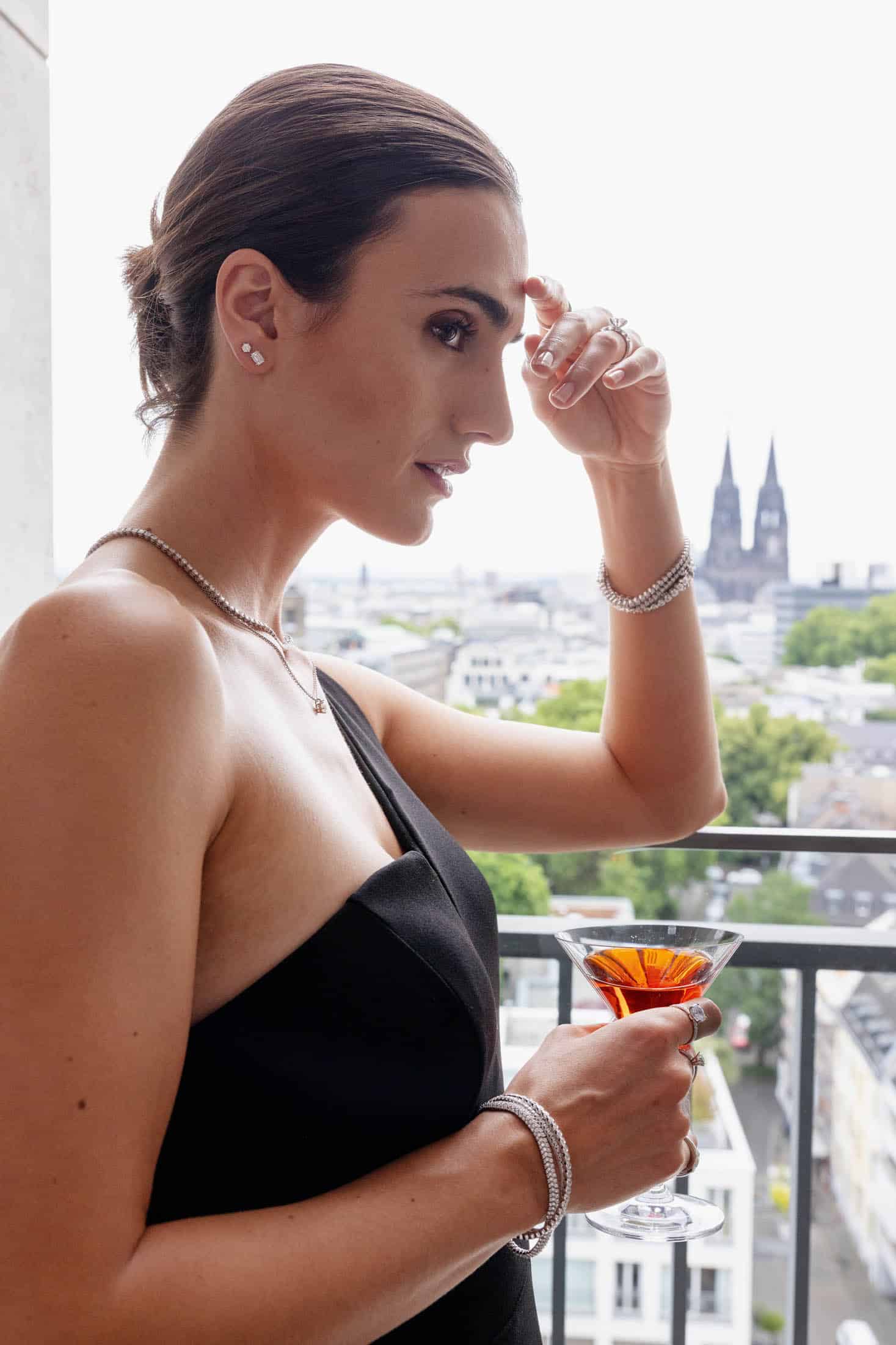 Eine Frau in einem eleganten schwarzen Kleid steht auf einem Balkon mit Blick auf die Stadtlandschaft und hält einen Cocktail in der Hand; im Hintergrund ist eine Kathedrale zu sehen. Sie trägt Schmuck und blickt nachdenklich in die Ferne. © Fotografie Tomas Rodriguez