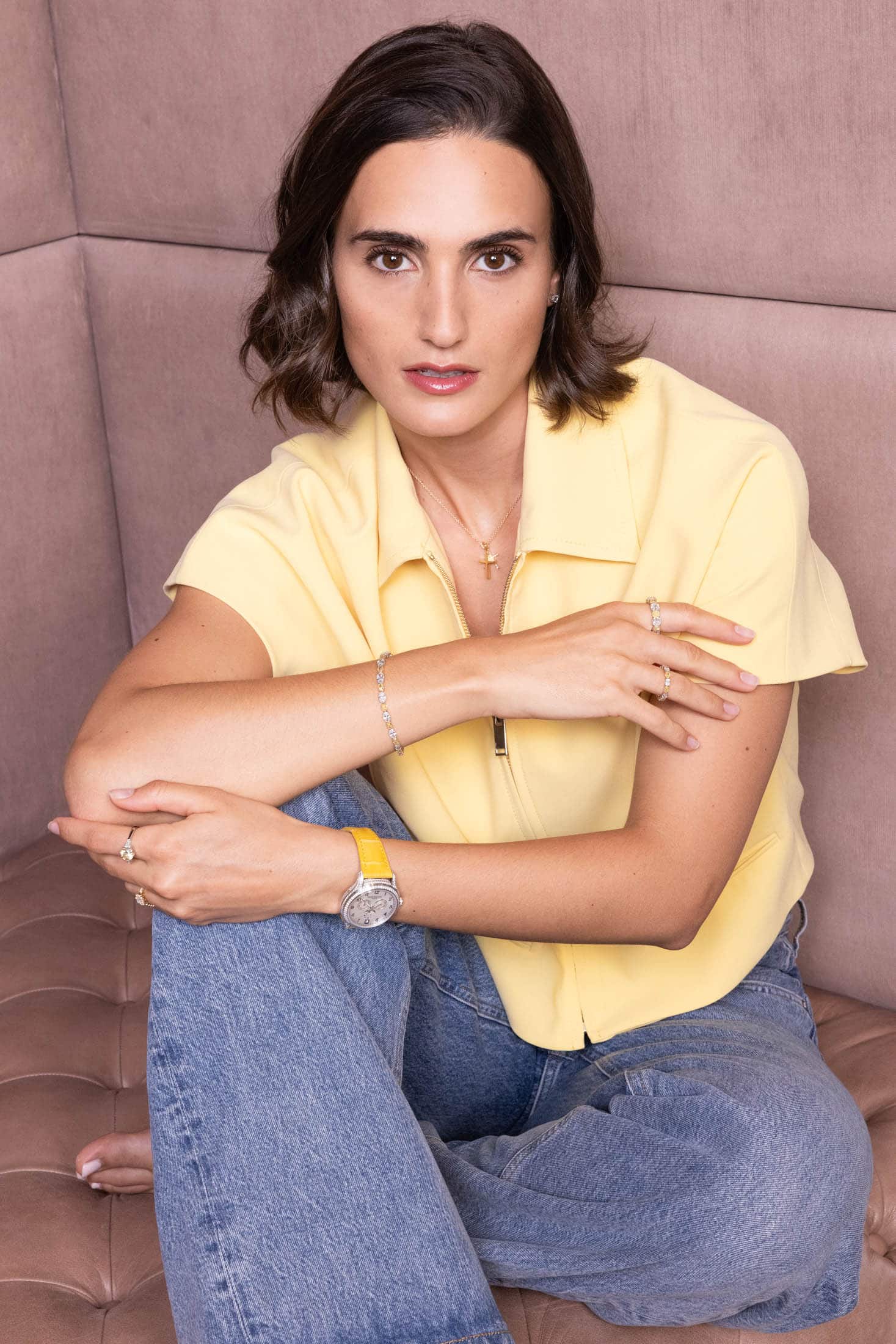 Eine Frau mit kurzen braunen Haaren sitzt mit gekreuzten Beinen auf einer beigen, gepolsterten Bank. Sie trägt ein gelbes Kurzarm-Shirt, blaue Jeans und eine Armbanduhr sowie Schmuck und blickt mit entspanntem Gesichtsausdruck in die Kamera. © Fotografie Tomas Rodriguez