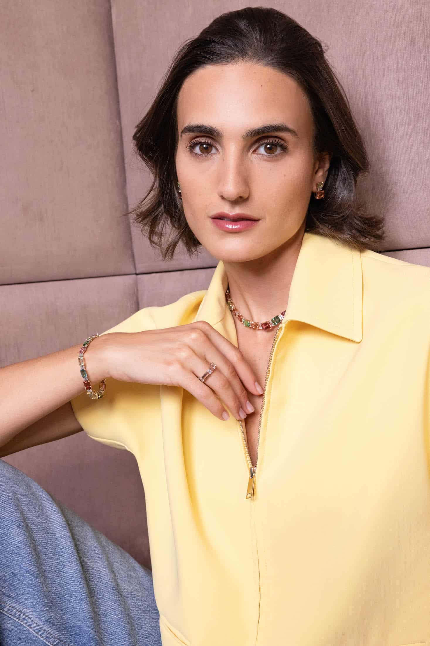 Eine Frau mit kurzen braunen Haaren trägt ein gelbes Shirt mit Reißverschluss, hellblaue Jeans und bunten Schmuck. Sie sitzt an einer gepolsterten beigen Wand und blickt mit entspanntem Gesichtsausdruck in die Kamera. © Fotografie Tomas Rodriguez