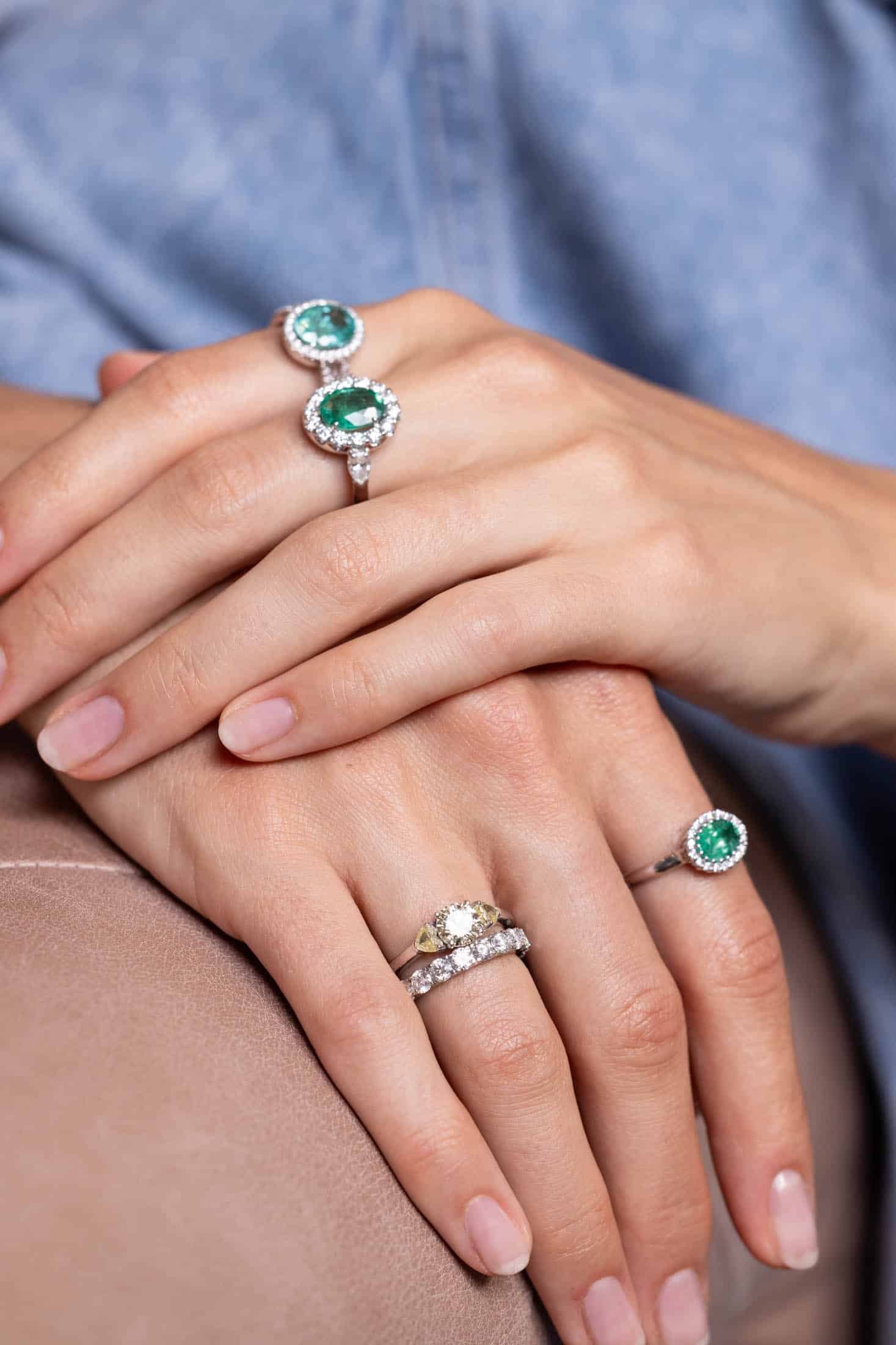 Eine Nahaufnahme von Händen, an denen Ringe mit grünen Edelsteinen und Diamanten stecken und die auf einer beigen Oberfläche ruhen. Die Person hat gepflegte Fingernägel und trägt ein blaues Hemd. © Fotografie Tomas Rodriguez
